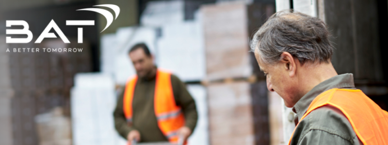 A man in the foreground, wearing a hi vis vest, looks at a tablet with data on it - while a another man in hi vis wheels a heavy-duty trolley laden with boxes towards the back of a heavy goods vehicle. British American Tobacco logo is in the top left corner.