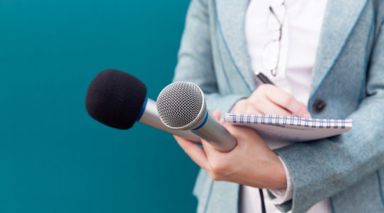 A journalist holding two microphones while writing notes down in a notepad.